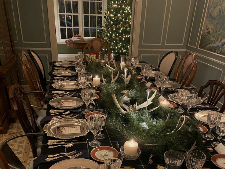 Uma mesa de Natal posta por Rafa: toalha xadrez escura, sousplats de prata, cristais, guirlanda de pinho com galhadas e velas, e a árvore de Natal acesa na janela.
